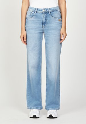 Lichtblauwe jeans met rechte pijpen, gecombineerd met witte sneakers en een wit shirt dat in de broek is gestopt, zichtbaar van taille tot voeten tegen een effen achtergrond.