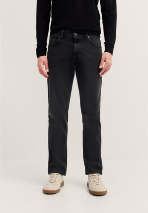 Mann in schwarzen Jeans, schwarzem Langarmoberteil und weißen Sneakers steht vor einem schlichten hellen Hintergrund.