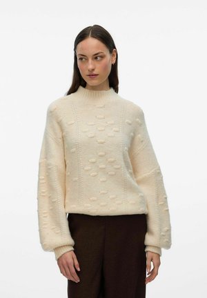 Maglione color crema con collo alto, caratterizzato da un motivo testurizzato, spalle scese e polsini a coste. Indossato con pantaloni marroni scuri.