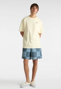 Helles gelbes Baumwoll-T-Shirt mit rundem Ausschnitt, kombiniert mit blauen karierten Badehosen. Die Hosen weisen auffällige quadratische Muster auf.