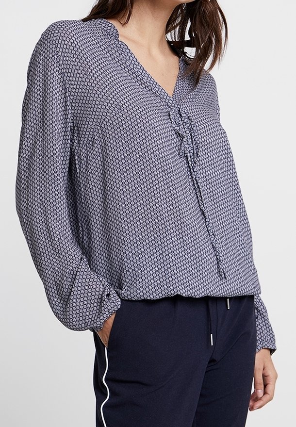 Femme portant un chemisier bleu à manches longues à motifs avec un col en V et un détail volanté, associé à un pantalon bleu marine avec une bande blanche sur le côté.