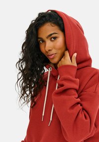 Červená mikina s kapucňou a nastaviteľnými šnúrkami, s rebrovanými manžetami a mäkkou textúrou. Kapucňa je položená dolu, odhaľujúc svetlú vnútornú podšívku.
