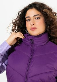 Lila Puffer-Weste mit hohem Kragen und Reißverschluss vorne. Glatte Textur mit gesteppten Details; abgebildet mit einem helllila Langarmshirt.