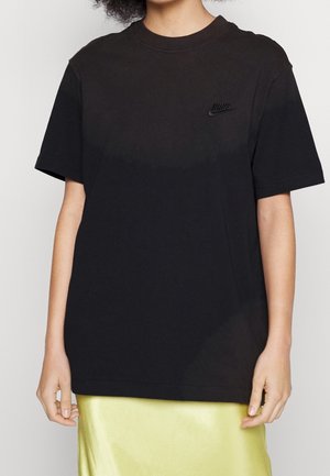 Person trägt ein schwarzes Nike-T-Shirt mit kurzen Ärmeln und kleinem Logo auf der Brust, kombiniert mit einem hellgrünen Satinrock.