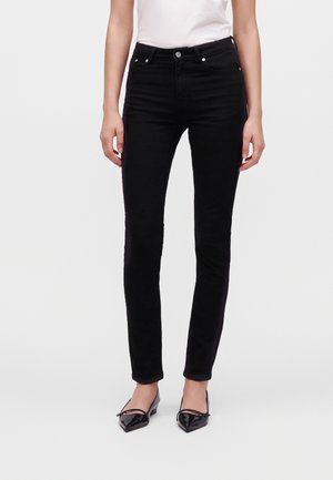 Femme portant un jean slim noir associé à des ballerines noires à bout pointu et un top blanc sans manches sur un fond uni.
