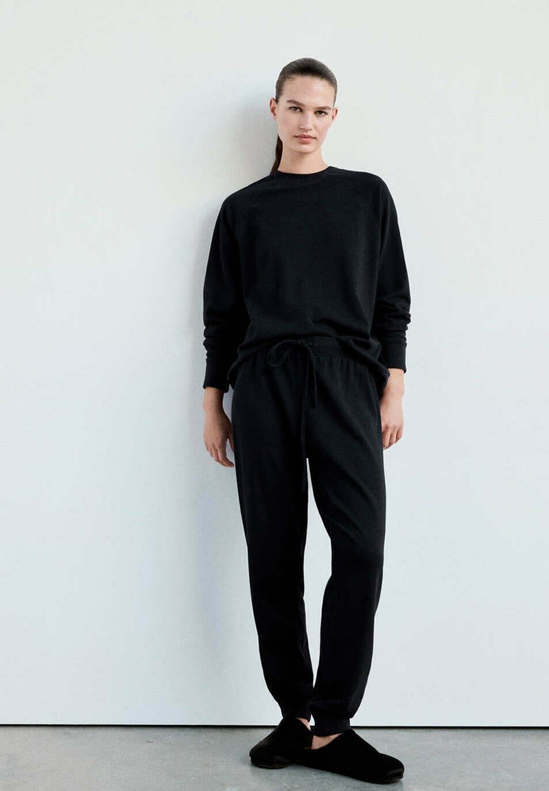 Femme debout contre un mur blanc portant un haut noir à manches longues, un pantalon noir avec cordon de serrage et des chaussures noires sans lacets.