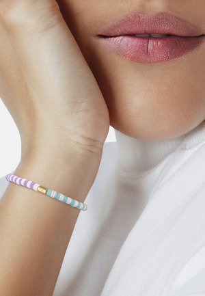 Primer plano de la parte inferior del rostro y la muñeca de una persona que lleva una pulsera de rayas en tonos pastel, apoyando la mano en la mejilla.