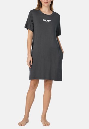 Frau steht barfuß und trägt ein lockeres dunkelgraues Kleid mit kurzen Ärmeln und dem "DKNY"-Logo auf der Brust sowie die Hände in den Taschen.