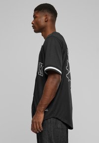 Maillot de baseball noir à manches courtes avec des accents blancs, présentant un graphique au dos et un ourlet légèrement incurvé en bas.