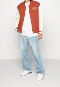 Röd och creamfärgad varsityjacka med vit broderi, tillsammans med en vit T-shirt, ljusblå jeans och vita sneakers. Avslappnad stil.