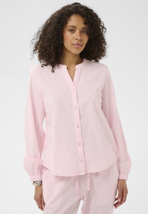 Femme aux cheveux bouclés portant une chemise à manches longues rose pâle boutonnée et un pantalon assorti, debout devant un fond uni.