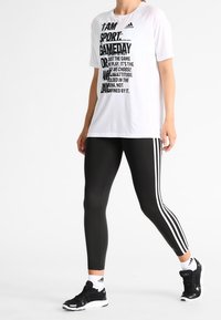 Bílé tričko s černým nápisem a logem 'adidas', doplněné černými legínami s třemi bílými pruhy po stranách a černými sportovními botami.