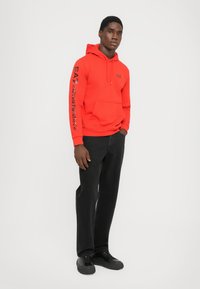 Sudadera roja de mezcla de algodón con bolsillo frontal, que presenta el logo negro de EA7 en la parte delantera y las mangas. Combinada con vaqueros y zapatos negros.