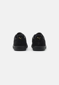 Schwarze Wildleder-Sneaker mit einer strukturierten Gummi-Sohle, versehen mit goldenen Puma-Logo-Akzenten an der Ferse und an der Seite sowie einem Low-Top-Design.