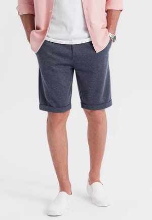 Navy blauwe shorts met een geweven textuur, opgerolde manchetjes en zijzakken, gedragen met een lichtroze shirt en witte instapschoenen.