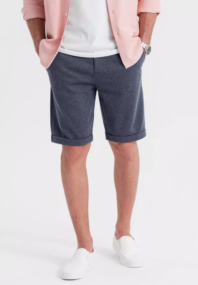 Shorts bleu marine avec une texture tissée, des revers roulés et des poches latérales, portés avec une chemise rose clair et des chaussures blanches à enfiler.