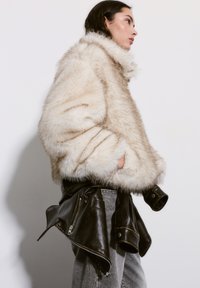 Faux fur jacket in beige with a high collar; features a textured surface. Paired with a black leather jacket tied at the waist and gray jeans.