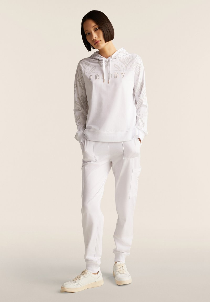 Sudadera blanca con "VIERNES" bordado en rosa claro, con mangas estampadas. Combinada con pantalones deportivos blancos y zapatillas crema.