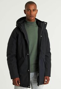 Crna vodootporna jakna s kapuljačom, prednjim zipom i kopčama na dugmad. Ima dva bočna džepa i meki zeleni pulover s rol kragnom ispod.