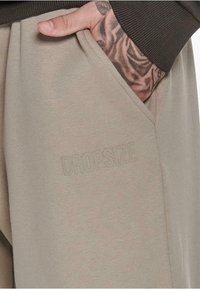 Beige sweatpants zijn voorzien van een zijzak met een subtiele geëtste "DROPSIZE" logo, gemaakt van een zachte, gestructureerde stof met een losse pasvorm.