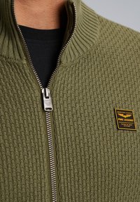 Olivgrüner Zip-Pullover mit strukturiertem Strickmuster, ausgestattet mit einem Metallreißverschluss und einem Markenlogo auf der Brust.