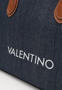Denim toteväska med bruna läderdetaljer. Har vit "VALENTINO"-logotyp, slät yta och en strukturerad form.
