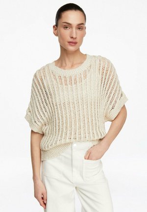 Woman wearing a beige open-knit short-sleeve sweater and high-waisted off-white pants with one hand in pocket.