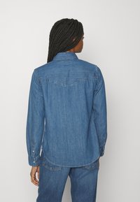 Denimhemd in mittlerem Blau, langärmlig mit Knopfmanschetten, spitzem Kragen und abgerundetem Saum. Mit Rückenteil-Details und strukturiertem Stoff.