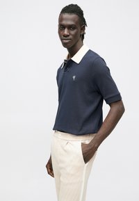 Marineblå pikéskjorte med beige krage, teksturert stoff, brodert logo på brystet. Kombinert med lyserosa mønstrede bukser.