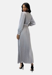 Lungo abito grigio plissettato con corpo aderente, maniche a sbuffo e una cintura. La vista posteriore mostra una texture liscia e un'elegante silhouette. Tacchi neri.