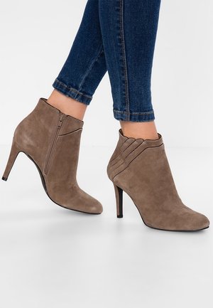 Bottes stilettos en daim taupe montant à la cheville avec fermeture éclair latérale, portées avec un jean bleu foncé retroussé.
