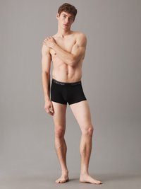 Fekete pamut boxer alsónadrág széles elasztikus derekának cégnévvel. A modell mezítláb áll egy semleges szürke háttér előtt.