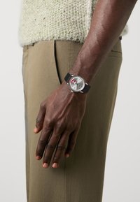 Montre analogique en argent avec un bracelet en caoutchouc noir, présentant un boîtier rond, des accents blancs et rouges, et un cadran texturé. Portée au poignet d'un homme.