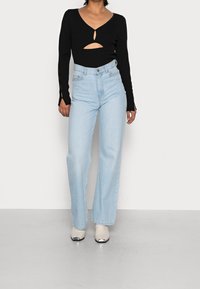 Top en tricot noir avec des découpes, associé à un jean large bleu clair taille haute. Le mannequin porte des bottes à talons beiges avec des accents métalliques.
