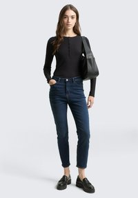 Naine kannab musta pikka varrukaga särk, tumesiniseid teksasid, musti nahast mokasíne ning hoiab käes suurt musta nahast õlakotti.