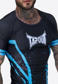 Melnā sporta krekls ar ziliem akcentiem un traipu dizainu. Ir TAPOUT logo, pieguļošs stils un kontrastējoša šuvēšana gar malām.