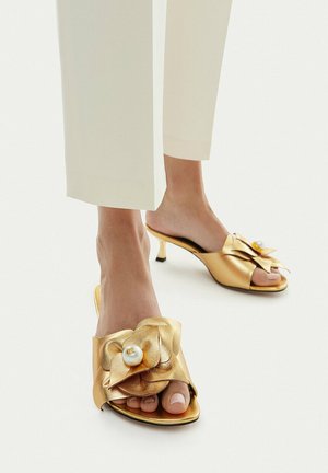 Pies con mules dorados de tacón kitten abierto en la punta, con gran detalle floral y adornos de perlas, combinados con pantalones blancos recortados.