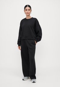 Jovem mulher com um moletom preto e calças largas pretas, de pé com uma mão no bolso, usando ténis cinzentos e brancos e brincos de argola.