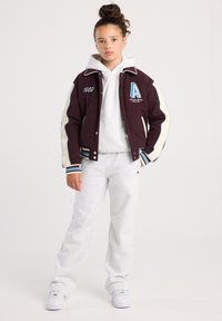 Maroon varsity jacket with cream accents, embroidered “A” and year "1989," paired with a gray hoodie and sweatpants. White sneakers.