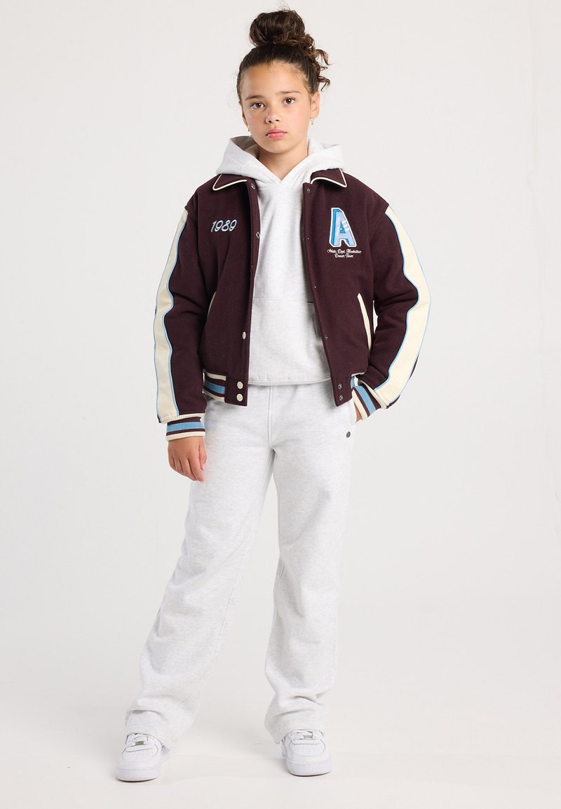 Junge Person steht mit Händen in den Taschen, trägt eine dunkle Collegejacke über einem weißen Kapuzenpullover, weiße Jogginghose und weiße Sneakers.