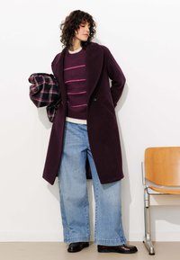 Long manteau en laine violet sur un pull à rayures bordeaux, associé à un jean large bleu clair et des chaussures noires cirées. Écharpe à carreaux à la main.