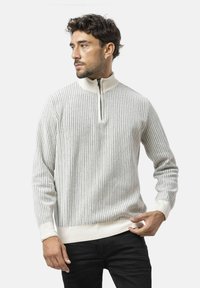 Hellgrauer, strukturierter Pullover mit vertikalen Rippen, Kragen mit halbem Reißverschluss, cremefarbigen Akzenten und körperbetontem Schnitt, getragen über dunklen Hosen.