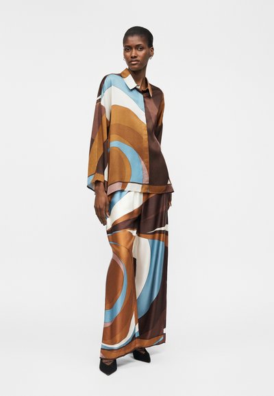 Nainen seisoo yllään löysä, pitkähihaista paitaa ja leveälahkeiset housut, joissa on abstrakteja ruskeita, sinisiä, valkoisia ja hiekansävyisiä kiemurakuvioita.