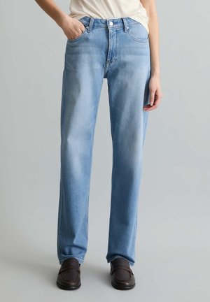 Person, die hellblaue Jeans mit geradem Bein, weißes Oberteil und dunkelbraune Slipper trägt, steht vor einem schlichten grauen Hintergrund.