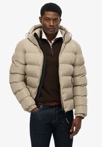 Beige pufferjack met een capuchon, voorzien van een ritssluiting aan de voorkant, zwarte voering en een geborduurd ontwerp. Draagt over een bruine trui en blauwe jeans.