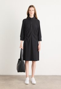 Zwarte jurk met hoge kraag, kwart-rits sluiting, geplooide zijkanten en driekwartmouwen. Gestyled met een zwarte handtas en witte sneakers.