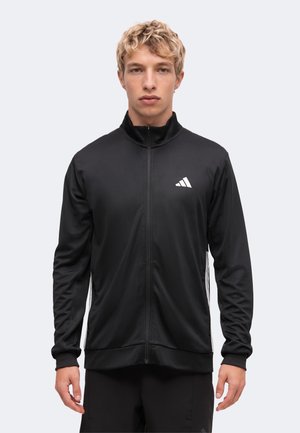 Joven con cabello rubio rizado que lleva una chaqueta deportiva negra con cremallera y un logo blanco en el pecho, de pie frente a un fondo claro y liso.