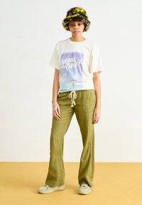 T-shirt crème à manches courtes avec impression graphique, pantalons en lin larges verts et un chapeau seau texturé et coloré. Des baskets neutres complètent la tenue.