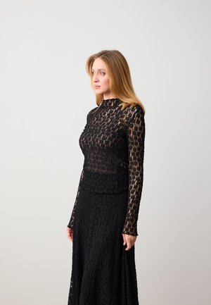 Femme blonde portant un haut noir à manches longues en dentelle et une longue jupe assortie, debout devant un fond gris, regardant sur le côté.