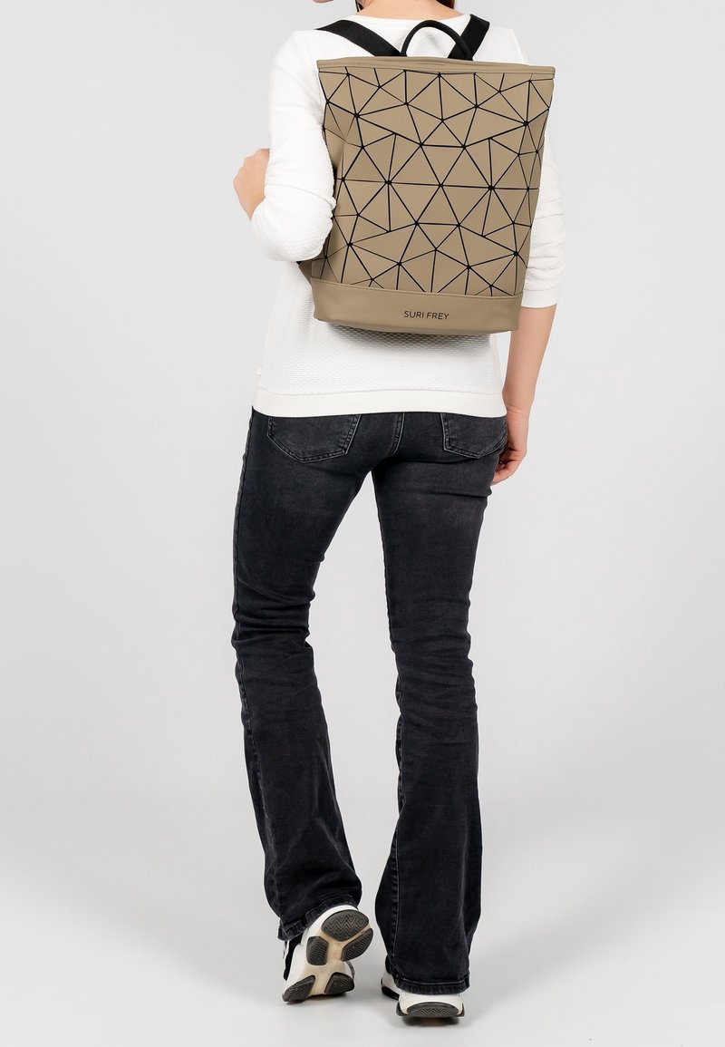 Personne portant un sac à dos beige à motifs géométriques avec l'étiquette "SURI FREY", un top à manches longues blanc, un jean noir et des baskets noir et blanc.